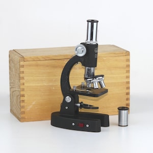 Könnte beinhalten: Ein schwarzes Mikroskop mit einer Holzkiste und einem silbernen Rohr. Das Mikroskop hat einen silbernen Knopf und eine silberne Linse. Die Kiste ist aus Holz und hat einen Klappdeckel.