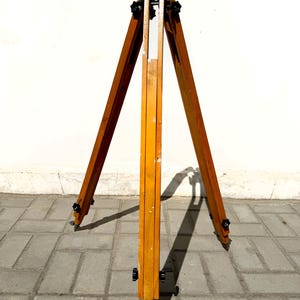 May include: A wooden tripod stand featuring three adjustable legs with black plastic knobs for securing height. The light brown wood has a smooth finish, and the structure is designed to support equipment with a central mounting point at the top.