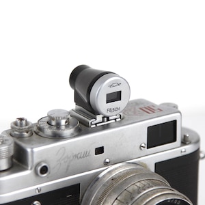 Puede incluir: Una cámara de película de 35 mm de color plateado y negro con un fotómetro incorporado y una lente con una apertura máxima de f/8,5 cm. La cámara tiene un telémetro y un visor.