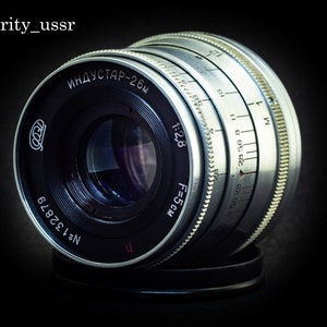 以下が含まれることがあります： 銀と黒のカメラレンズで、側面に「ИНДУСТАР-26м」と「№1 32879」の文字が印刷されています。レンズには被写界深度スケールとフォーカスリングがあります。