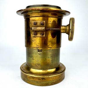 May include: A vintage brass camera lens with a circular top and base. The lens has a cylindrical body with a knob on the side and several screws. The brass has a worn, aged appearance.
