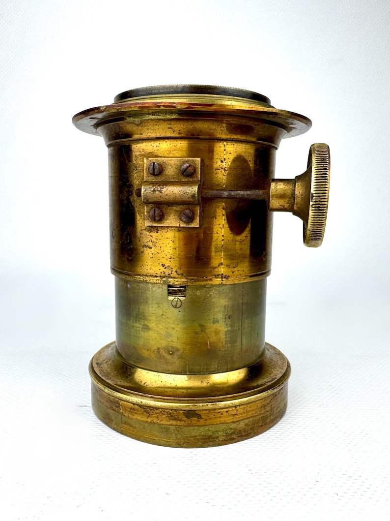 May include: A vintage brass camera lens with a circular top and base. The lens has a cylindrical body with a knob on the side and several screws. The brass has a worn, aged appearance.