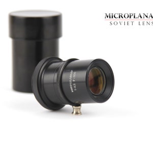 Puede incluir: Un objetivo soviético MicroPlanar negro con el texto "Микропланар 1:4.5 F=100" impreso en el lateral. El objetivo está enfocado y la apertura está abierta.