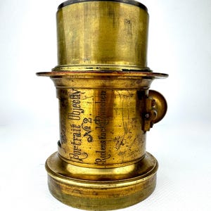 May include: A vintage brass camera lens with the text "Portrait Objectiv No. 2" and "Rodens tock Munchen" etched on the side. The lens has a cylindrical shape with a flared base and a top section. The brass has a worn, aged appearance.