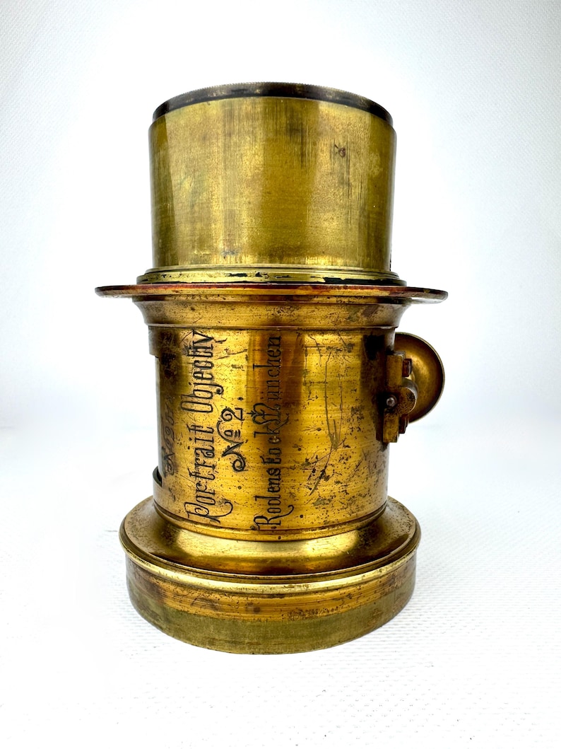 May include: A vintage brass camera lens with the text "Portrait Objectiv No. 2" and "Rodens tock Munchen" etched on the side. The lens has a cylindrical shape with a flared base and a top section. The brass has a worn, aged appearance.