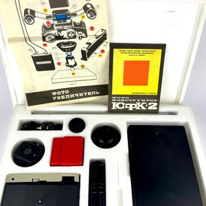 Puede incluir: Un kit de ampliadora de fotos vintage, que incluye varios componentes negros y rojos, un diagrama y un folleto. El kit está alojado en un inserto de espuma blanca, con el texto "ФОТО УВЕЛИЧИТЕЛЬ" visible en el diagrama.