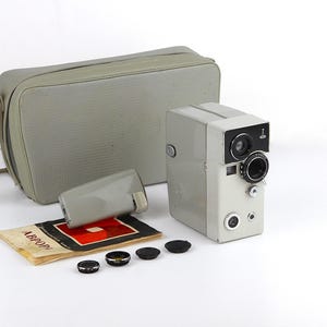 May include: A vintage gray film camera with a matching case, flash, and several lens filters. The camera has a black lens and control dials. A booklet with red and black accents is also included, along with a gray carrying case.