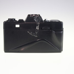 May include: A black 35mm film camera with a built-in flash. The camera has a few scratches and scuffs on the surface.