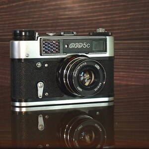 May include: A black and silver 35mm film camera with a lens labeled "Industar-61 L/M 4/55". The camera has a silver top plate with the text "ФЭД 5С" printed on it.