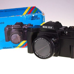 May include: A black 35mm manual camera with a lens labeled "New Color Optic" and "Belka U.S.A." The camera is in its original box, which is blue with yellow and red stripes and the text "Keep The Memories Forever" and "Image Master".
