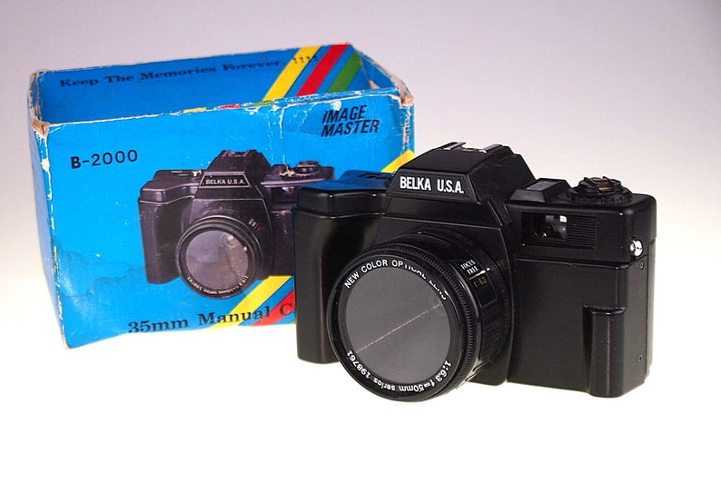 May include: A black 35mm manual camera with a lens labeled "New Color Optic" and "Belka U.S.A." The camera is in its original box, which is blue with yellow and red stripes and the text "Keep The Memories Forever" and "Image Master".