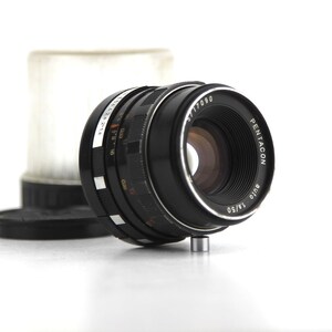 May include: A black Pentacon Auto 1/50 lens with a silver ring and markings. The lens is designed for a 35mm camera.