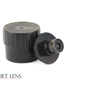 Puede incluir: Un objetivo de microscopio negro con el texto "МИКРОПТО" y "112.5" impreso en él. El objetivo está en una caja de plástico negro.