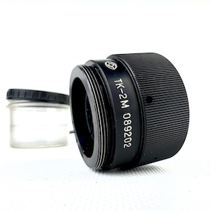 Puede incluir: Un adaptador de lente de cámara negro con el texto "TK-2M 089202" impreso en el lateral. El adaptador tiene un exterior estriado y un interior roscado. Un pequeño tarro de cristal con tapa negra está en el fondo.