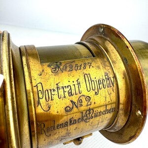 May include: Close-up of a vintage brass Portrait Objectiv No. 2 lens. The lens has the number 25187 etched on it, along with the words "Portrait Objectiv No 2" and "Rodens Stock M&uuml;nchen" in elegant script. The brass has a warm, aged patina.
