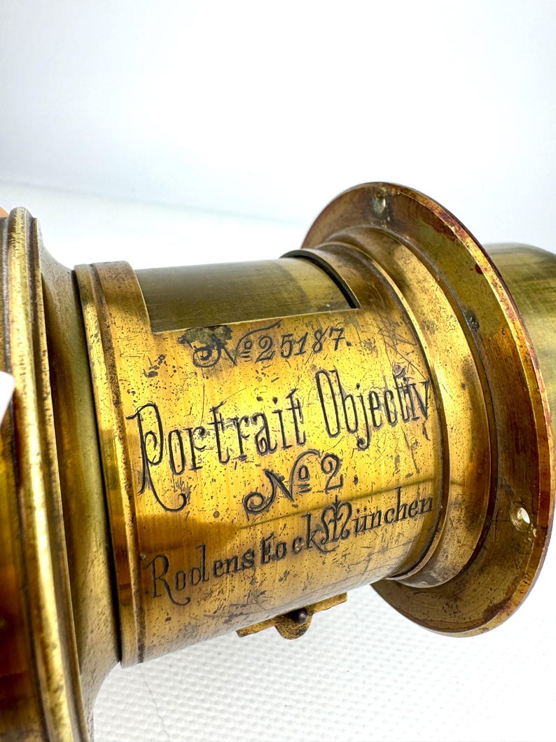 May include: Close-up of a vintage brass Portrait Objectiv No. 2 lens. The lens has the number 25187 etched on it, along with the words "Portrait Objectiv No 2" and "Rodens Stock M&uuml;nchen" in elegant script. The brass has a warm, aged patina.