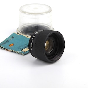 Puede incluir: Un objetivo de cámara negro con el texto "TK-2M 017273" se muestra sobre un fondo blanco. También hay un recipiente de plástico transparente y un papel azul y verde. El objetivo tiene una rosca frontal.