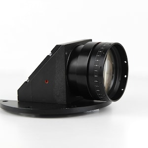 May include: A black camera lens with a series of measurement markings and a clear glass lens. The lens is mounted on a black angled bracket and a curved base, set against a white background.