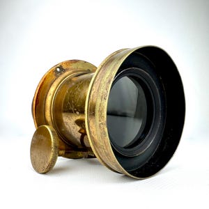 May include: Antique brass-colored lens with a black lens element. The lens has a circular shape with a textured, aged appearance. A small, round adjustment knob is visible on the side. The lens is designed for photography or optical use.