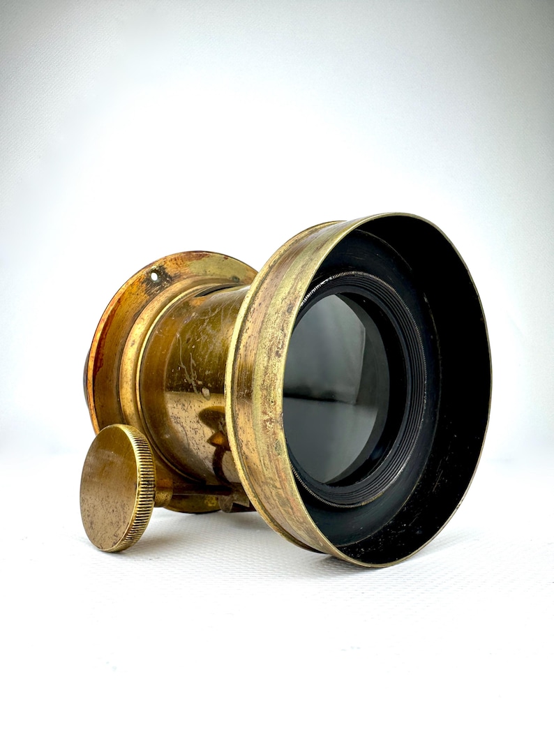 May include: Antique brass-colored lens with a black lens element. The lens has a circular shape with a textured, aged appearance. A small, round adjustment knob is visible on the side. The lens is designed for photography or optical use.