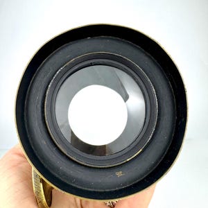 May include: Close-up of a vintage brass and black lens. The lens has a circular shape with a clear glass center and a black rubberized ring. The brass rim shows signs of age. The lens is held in a hand.