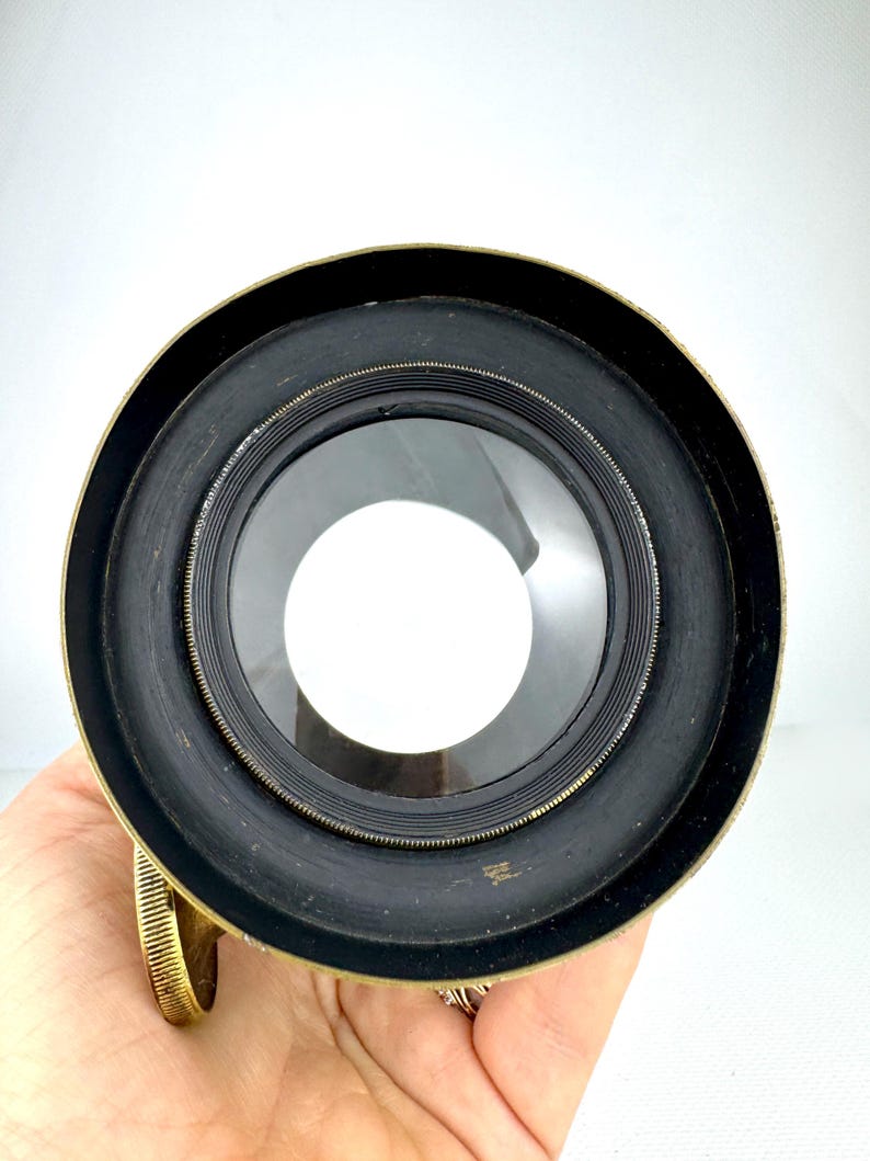 May include: Close-up of a vintage brass and black lens. The lens has a circular shape with a clear glass center and a black rubberized ring. The brass rim shows signs of age. The lens is held in a hand.