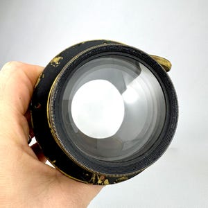 May include: A vintage camera lens with a black and gold finish. The lens has a large, clear glass element and a brass adjustment ring. The lens is held in a hand against a white background.
