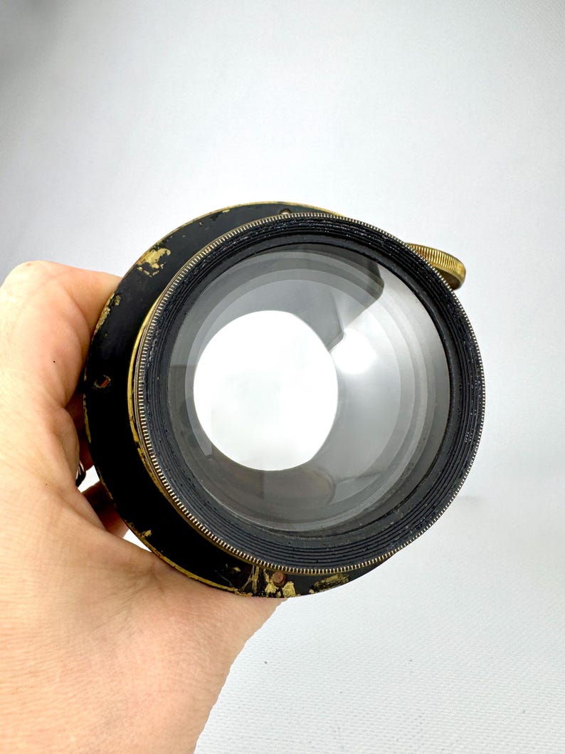 May include: A vintage camera lens with a black and gold finish. The lens has a large, clear glass element and a brass adjustment ring. The lens is held in a hand against a white background.