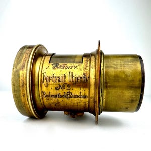May include: Antique brass camera lens with the text "Portrait Objectiv No. 2 Rodenstock M&uuml;nchen". The lens has a cylindrical shape with a series of rings and a flange. The brass has a worn, aged appearance.