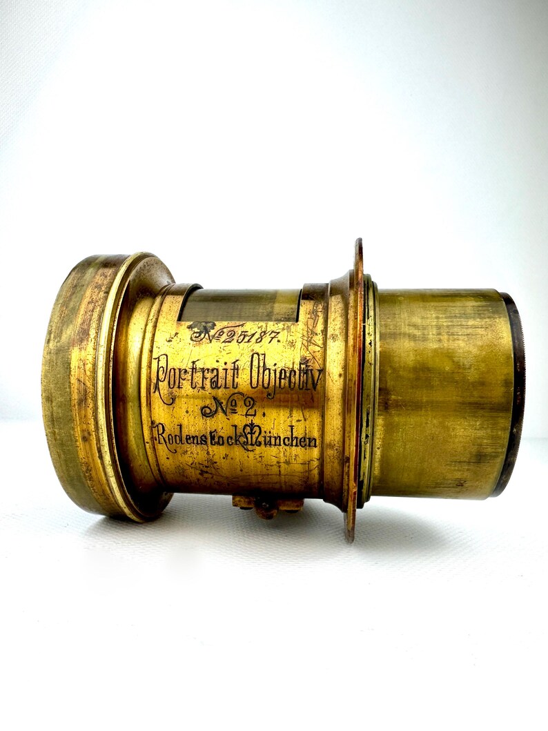 May include: Antique brass camera lens with the text "Portrait Objectiv No. 2 Rodenstock M&uuml;nchen". The lens has a cylindrical shape with a series of rings and a flange. The brass has a worn, aged appearance.