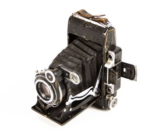 May include: A vintage black and silver folding camera with a leather bellows and a lens with a silver ring. The camera has a leather strap attached to the top.