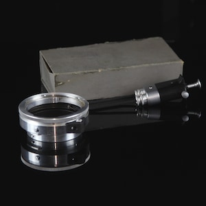 Op de afbeelding: Een zilveren camera-objectiefadapterring met een zwarte knop en een zwarte en zilveren camera-objectiefverlenging.