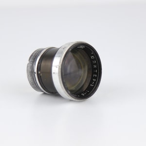 Può includere: Un obiettivo per fotocamera vintage con corpo nero e argento. L'obiettivo ha un elemento in vetro trasparente ed è etichettato con testo in lingua straniera. L'obiettivo è progettato per una fotocamera.