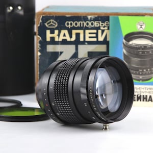 Op de afbeelding: Een zwarte camera lens met een zilveren ring en een zwarte lensdop. De lens is gemarkeerd met "T" en "Heliar-3". De lens ligt op een wit oppervlak met een doos en een lenshoes erachter.