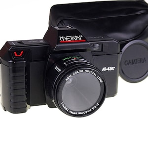 Puede incluir: Una cámara negra Meikai AR-4367 con una tapa de lente negra y una funda de cámara negra. La cámara tiene una lente negra con el texto "New Color Optical Lens 1:1.9 f=50mm 197861" impreso en ella.