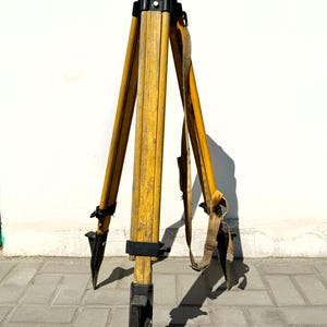 May include: A vintage surveyor tripod featuring three yellow wooden legs with black metal pointed feet and a black mounting head. The tripod includes a tan fabric carrying strap attached to the frame, showing signs of wear and weathered wood grain.