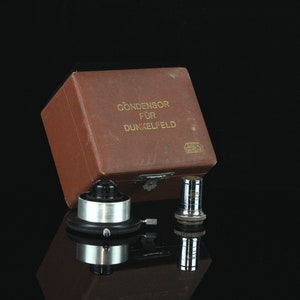 May include: A brown box with gold lettering that reads "Condensor für Dunkelfeld" and a small silver microscope condenser with a black base.