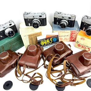 May include: A collection of vintage black and silver film cameras with leather cases, lens caps, and original packaging. The cameras are arranged with various documents and boxes, showcasing a retro aesthetic.