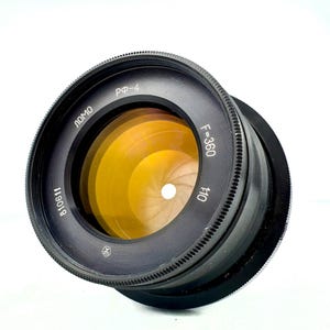 Op de afbeelding: Een zwarte LOMO RF-4 cameralens met een opvallend amberkleurig glaselement en een zichtbaar diafragma met meerdere lamellen. De voorste ring bevat de witte tekst LOMO, RF-4, F=360, 1:10 en het serienummer 810611.