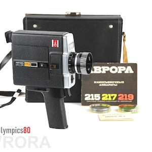 May include: A black and silver Avrora 215 movie camera with a black carrying case and a manual. The manual has the text "Avrora Kino-s'yemochnye apparaty 215 217 219".