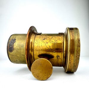 May include: A vintage brass camera lens with a detached circular cap. The lens has a cylindrical body with a series of rings and a slightly tarnished, aged appearance. The cap is also brass and shows signs of wear.