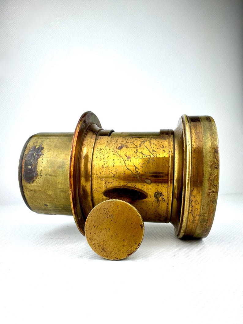 May include: A vintage brass camera lens with a detached circular cap. The lens has a cylindrical body with a series of rings and a slightly tarnished, aged appearance. The cap is also brass and shows signs of wear.