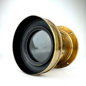 May include: A vintage brass lens with a black rim and a clear glass element. The lens has a circular shape and a weathered, antique appearance. The brass has a golden hue, and the lens is set against a white background.