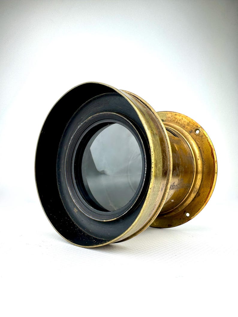 May include: A vintage brass lens with a black rim and a clear glass element. The lens has a circular shape and a weathered, antique appearance. The brass has a golden hue, and the lens is set against a white background.