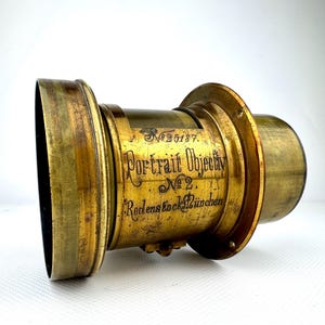 May include: Antique brass portrait lens with the text "Portrait Objectiv No. 2 Rodenstock M&uuml;nchen" and serial number 25187. The lens has a cylindrical shape with a flared front and a smaller rear section. The brass has a warm, aged patina.