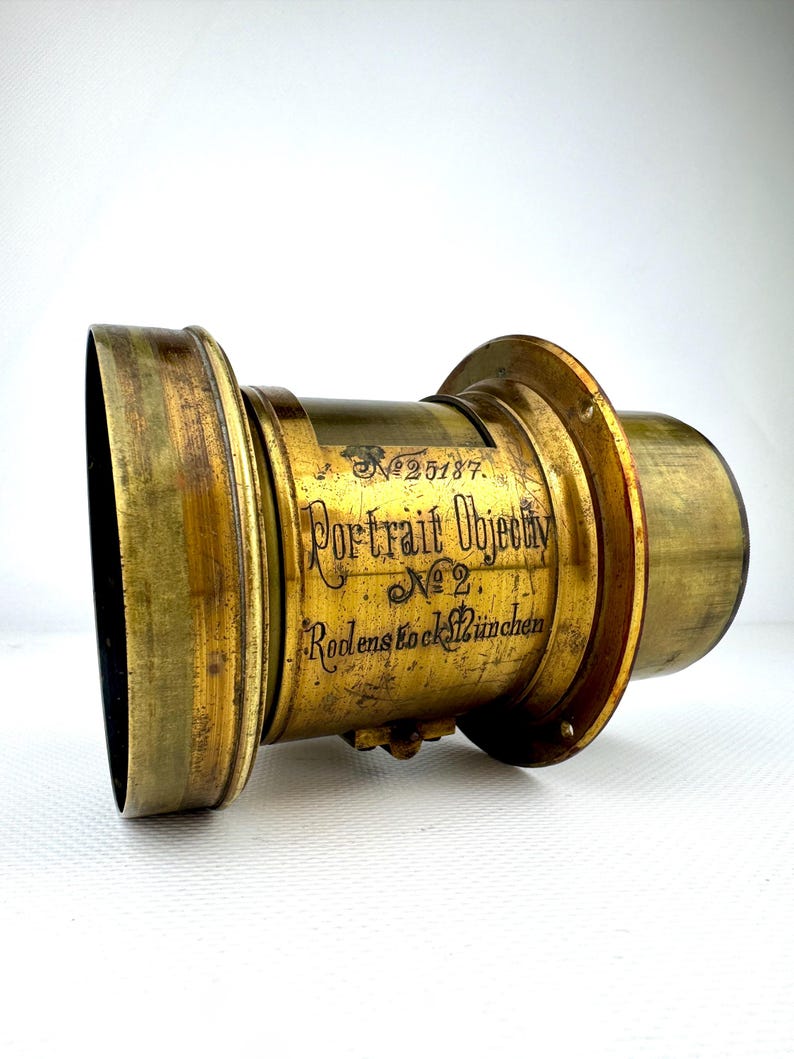 May include: Antique brass portrait lens with the text "Portrait Objectiv No. 2 Rodenstock M&uuml;nchen" and serial number 25187. The lens has a cylindrical shape with a flared front and a smaller rear section. The brass has a warm, aged patina.