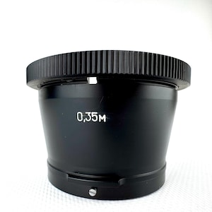 May include: A black camera lens with a ridged top and a tapered body. The lens has the number "0,35M" printed in white. The lens is designed for photography and is likely used with a camera.