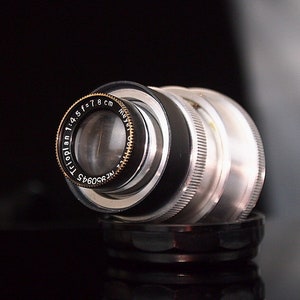 Puede incluir: Un objetivo de cámara plateado con detalles negros y un anillo dorado alrededor del elemento frontal. El objetivo está etiquetado como "Trioplan 1:4.5 f=7.8 cm Nr. 360945 Meyer Optik"