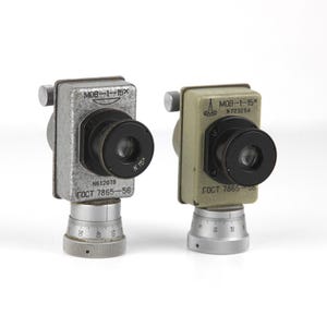 Puede incluir: Dos instrumentos ópticos de metal plateado y verde con lentes negras y un dial en la parte inferior. Los instrumentos están etiquetados como "MOB-1-15X" y tienen el texto "GOCT 7865-56" en ellos.