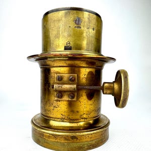 May include: Antique brass lamp component with a cylindrical body, a circular base, and a ridged knob. The brass has a tarnished, aged appearance, suggesting its vintage. The component features a series of screws and a small lever. The overall design is industrial.